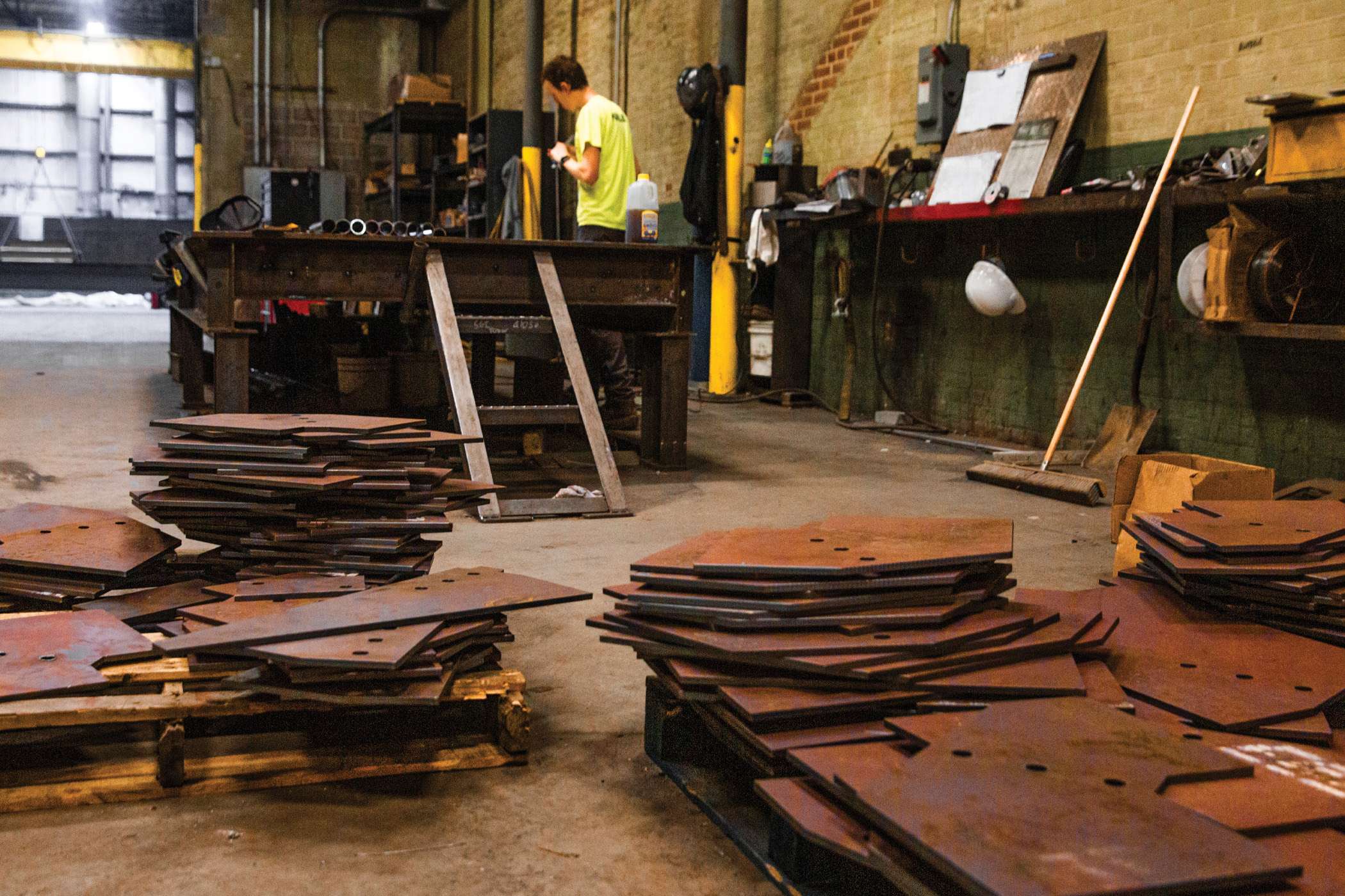 Steel plates are piled up in a warehouse with a man in a high visibility shirt standing in the background.