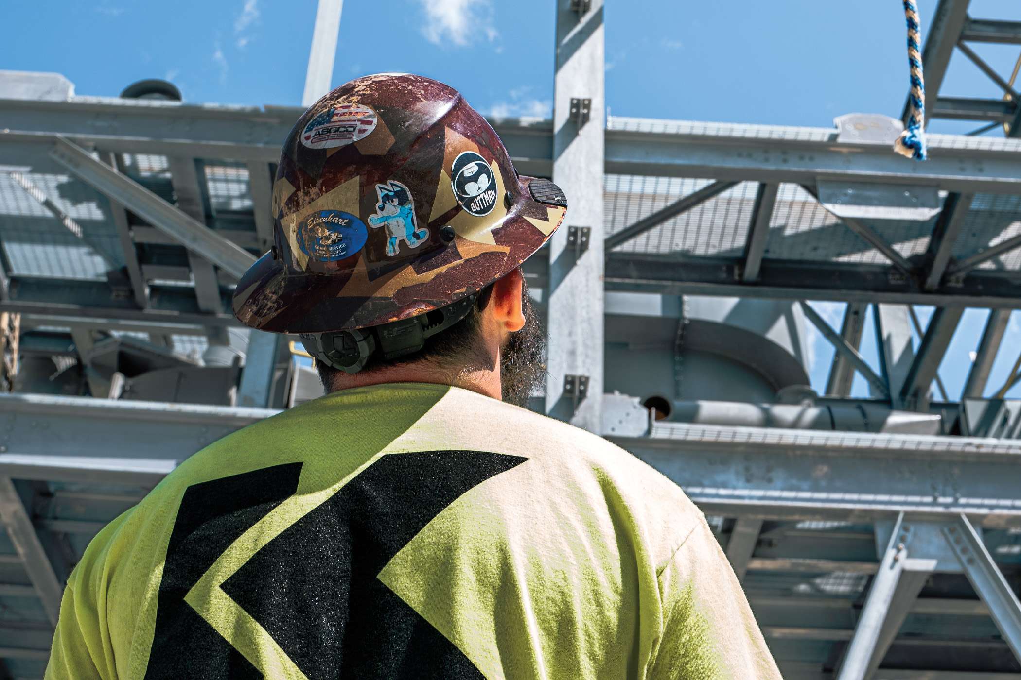A construction worker stands before a large metal structure wearing a hard hat adorned with stickers.