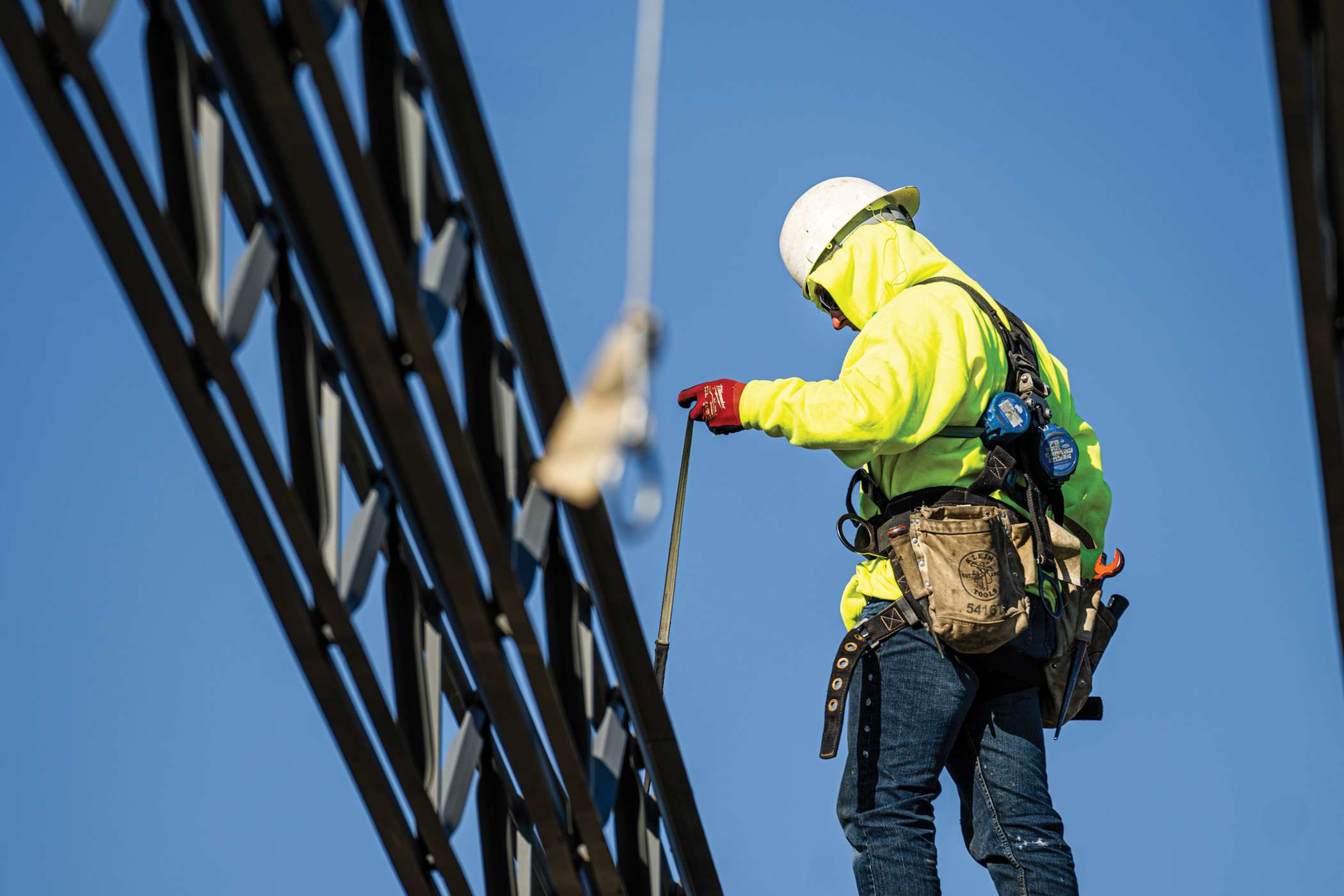 A construction worker in safety gear uses a tool while high on a building's framework.