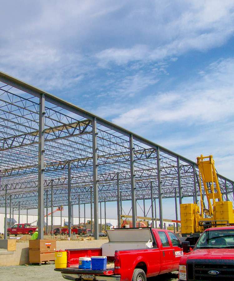 An unfinished steel frame building is under construction with trucks and equipment nearby.
