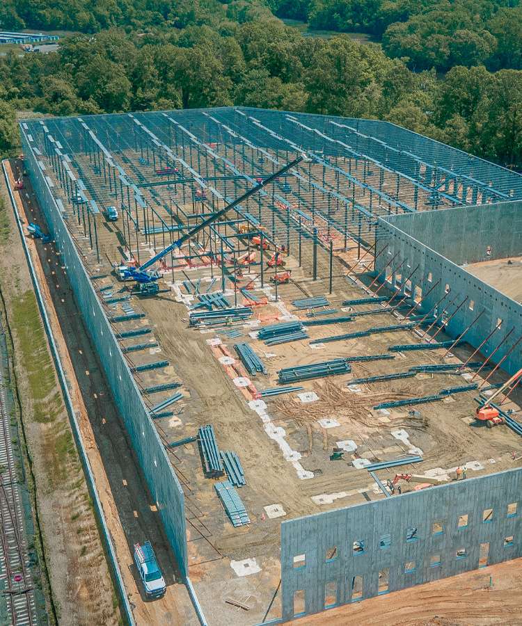 An aerial view shows a large, partially-constructed building with a steel frame and concrete walls, surrounded by construction equipment and materials.