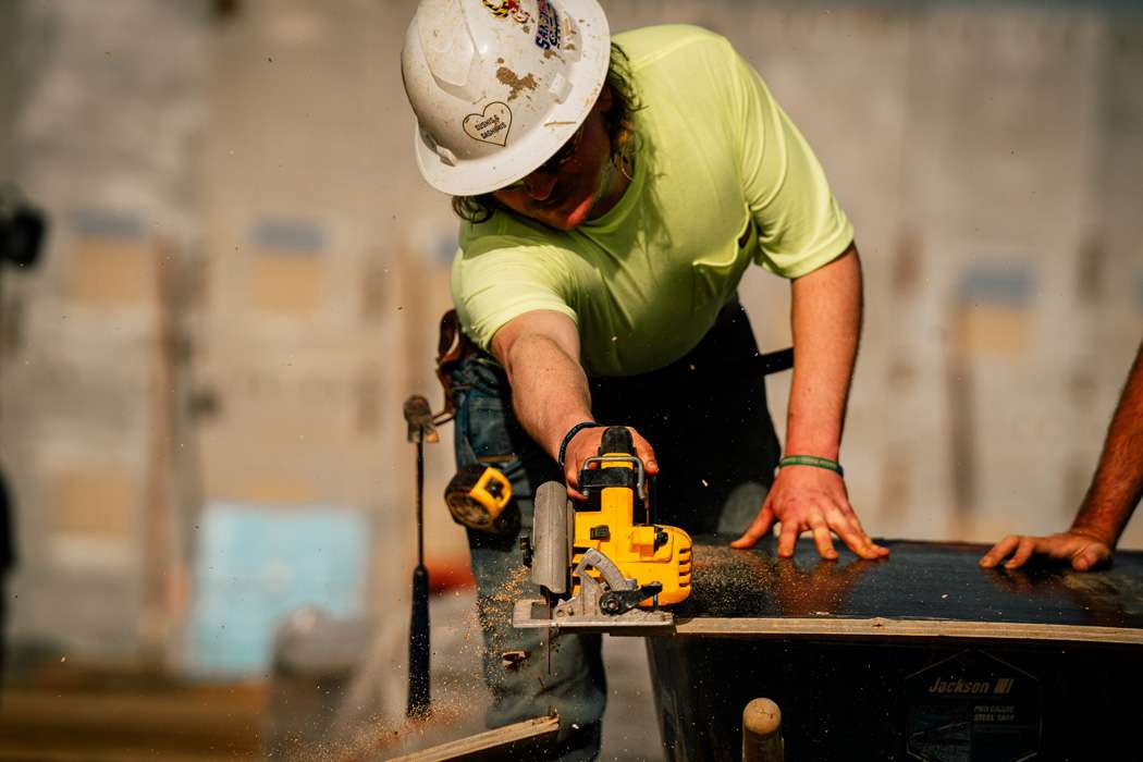 A construction worker wearing a hard hat and safety glasses cuts a piece of wood with a circular saw.