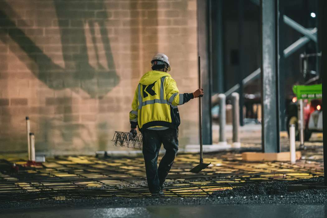 A construction worker in a yellow jacket walks across a rebar-covered ground, holding a tool in each hand.