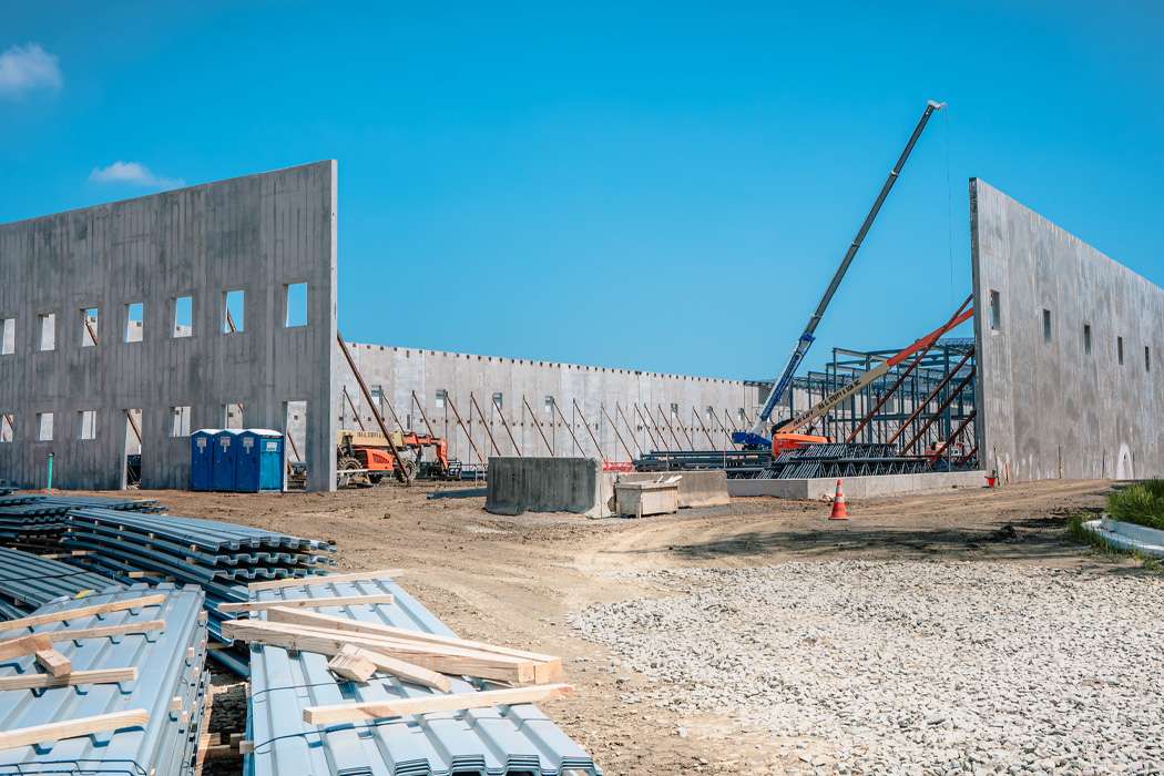 A construction site shows concrete walls being erected with the help of cranes and metal frameworks under a clear blue sky.