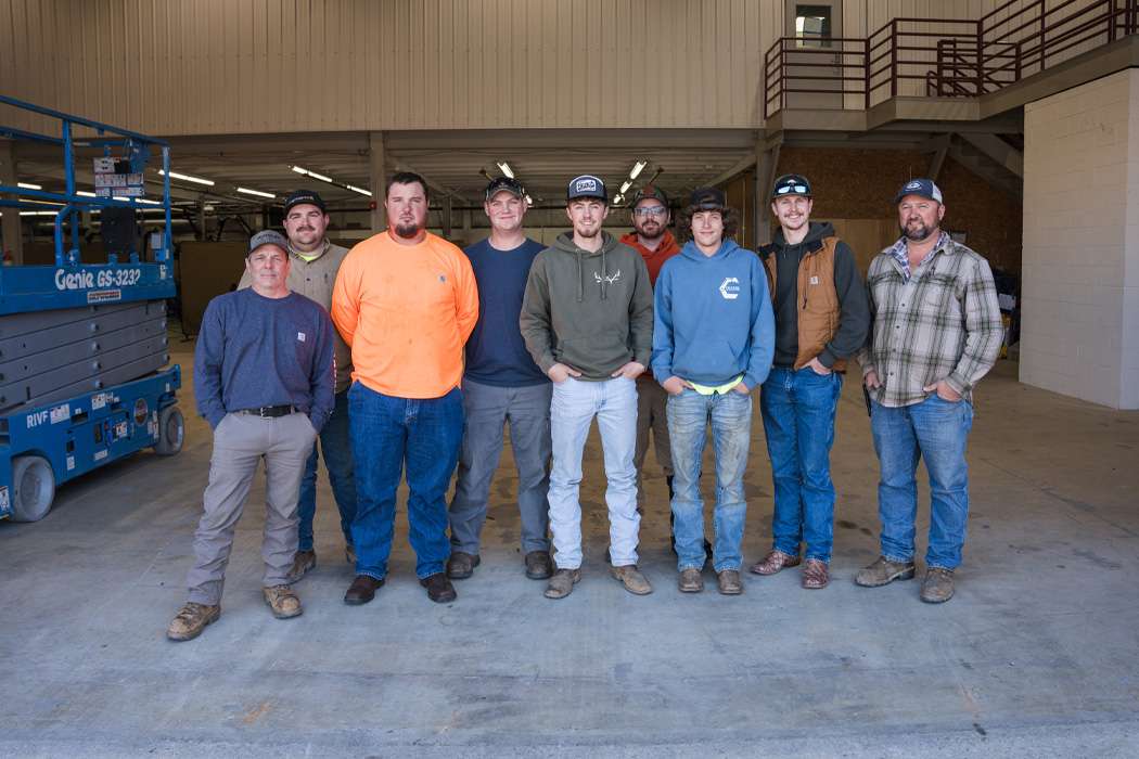 A group of nine construction workers, dressed in casual workwear and safety gear, stand together inside a large industrial building with concrete floors and equipment in the background.