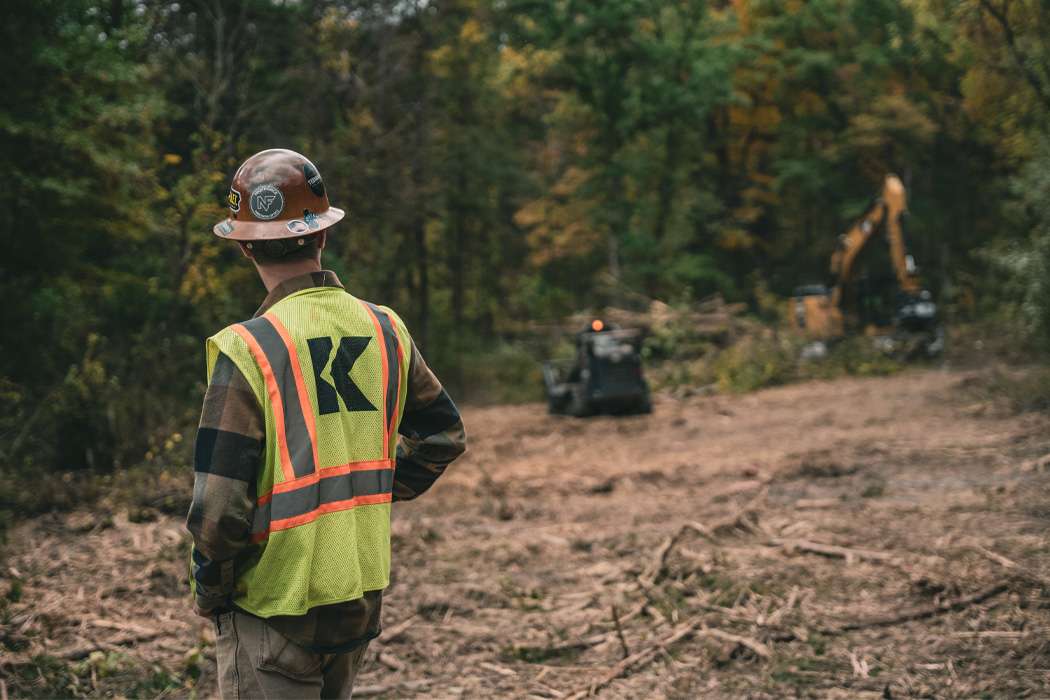 A construction worker in a hard hat and safety vest stands on a cleared patch of land, looking towards an excavator and another piece of equipment.