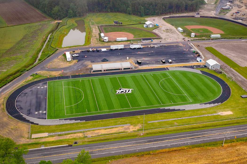 An aerial view showcases a sports complex with a football field, running track, baseball fields, and associated parking, set amid a landscape of trees and fields.