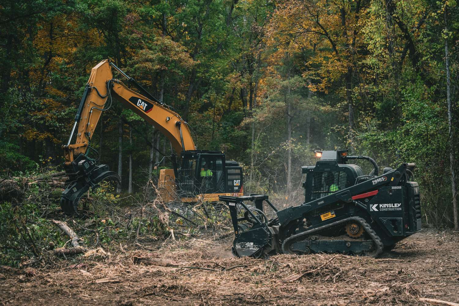 A yellow CAT excavator and black compact track loader clear trees and brush in a forested area.